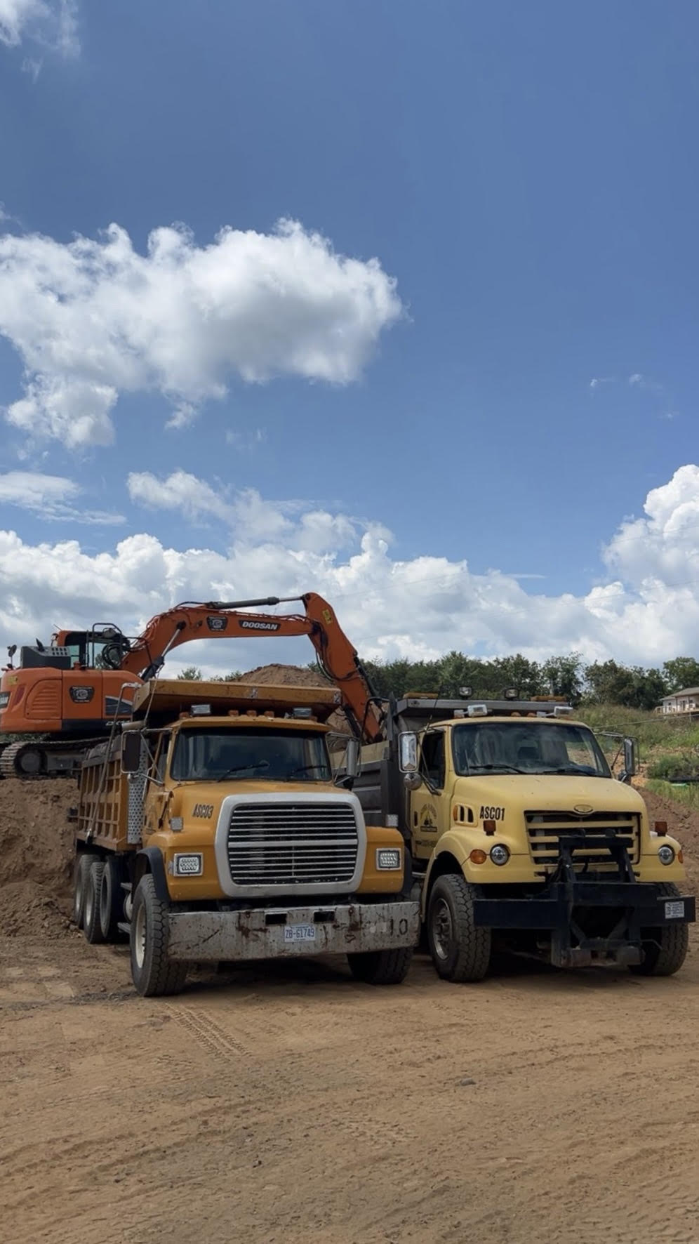 Two Angel & Sons dump trucks on site with an excavator loading dirt.